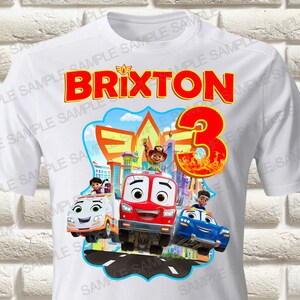 Pode incluir: Camiseta branca com um gráfico colorido de veículos de desenho animado do programa 'Super Wings'. O gráfico inclui o texto 'BRIXTON' e o número '3'.