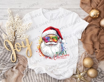 Puede incluir: Camiseta blanca con un gráfico colorido de Papá Noel con gafas de sol arcoíris y barba blanca. El texto "Feliz Navidad" está escrito debajo del gráfico.