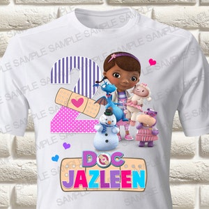 Puede incluir: Camiseta blanca con un número 2 morado y rosa, una venda con un corazón rosa y una imagen de dibujos animados de Doc McStuffins con sus amigos. El texto "Doc Jazleen" está debajo de la imagen.