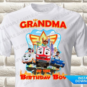 Op de afbeelding: Een wit t-shirt met korte mouwen en een kleurrijke afbeelding van cartoonvoertuigen en een personage. De tekst luidt GRANDMA OF THE BIRTHDAY BOY in dikke rode letters. Het ontwerp bevat een rode brandweerwagen, een blauwe auto en een ambulance.