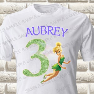 May include: White t-shirt featuring the name "Aubrey" in purple, a glittery green number "3", and a cartoon image of Tinkerbell in a green dress. The shirt is a children's birthday theme.