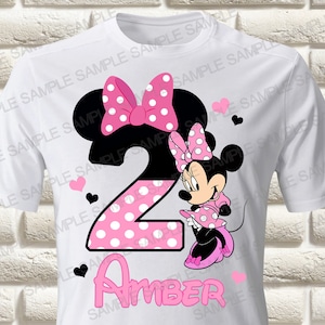 Könnte beinhalten: Weißes T-Shirt mit einem Minnie-Maus-Design für den zweiten Geburtstag. Das Design umfasst eine große Zahl 2 mit rosa Punkten, Minnie Maus, rosa Herzen und den Namen "Amber" in Rosa.