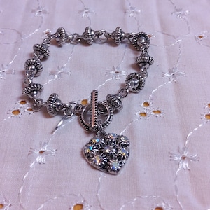 May include: A silver bracelet with a heart charm. The bracelet has a toggle clasp and is made of small silver beads. The heart charm is decorated with rhinestones.