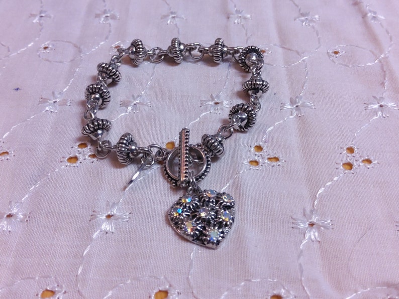 May include: A silver bracelet with a heart charm. The bracelet has a toggle clasp and is made of small silver beads. The heart charm is decorated with rhinestones.