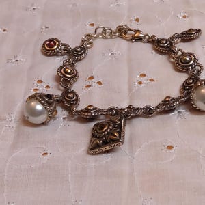 May include: Silver-toned charm bracelet featuring decorative links, pearl accents, and a dangling charm. The bracelet includes a mix of round and ornate elements, with a lobster clasp closure. The pearls are approximately 0.5 inches in diameter.