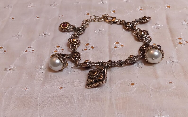 May include: Silver-toned charm bracelet featuring decorative links, pearl accents, and a dangling charm. The bracelet includes a mix of round and ornate elements, with a lobster clasp closure. The pearls are approximately 0.5 inches in diameter.