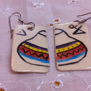 May include: A pair of rectangular earrings with a cream-colored base. Each earring features a hand-painted design of a stylized vessel with a black outline and colorful bands of red, yellow, blue, and purple. The earrings have silver-tone hooks.