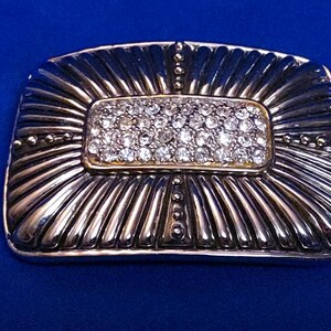 May include: Silver-toned rectangular belt buckle with a central rectangular section encrusted with small, clear crystals. The buckle has a sunburst design and is set against a blue background.