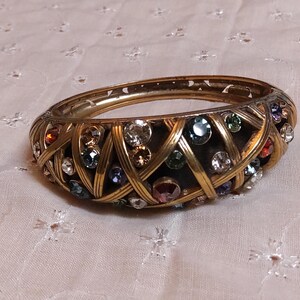 May include: A gold-toned bangle bracelet with a woven design. The bracelet is adorned with a variety of colorful gemstones, including clear, red, blue, and green. The bracelet is a vintage piece.
