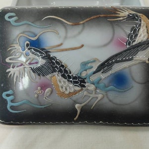 May include: A white porcelain box with a black lid. The lid features a hand-painted design of a black and gold dragon with blue and pink accents.