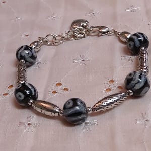 May include: A silver bracelet with black and white patterned beads and decorative silver accents. The bracelet features a heart-shaped charm and a clasp closure. The beads are round and oval-shaped, creating a unique jewelry piece.