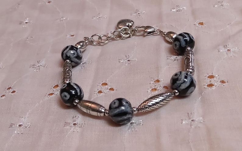 May include: A silver bracelet with black and white patterned beads and decorative silver accents. The bracelet features a heart-shaped charm and a clasp closure. The beads are round and oval-shaped, creating a unique jewelry piece.