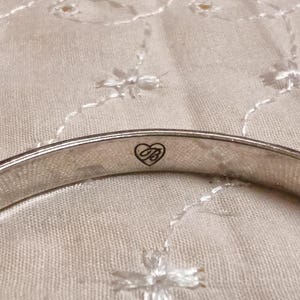 May include: Close-up of a silver-toned bangle bracelet. The bracelet has a smooth, polished surface and a heart-shaped logo with the letters "B" and "B" inside. The background is a cream-colored fabric with a floral pattern.