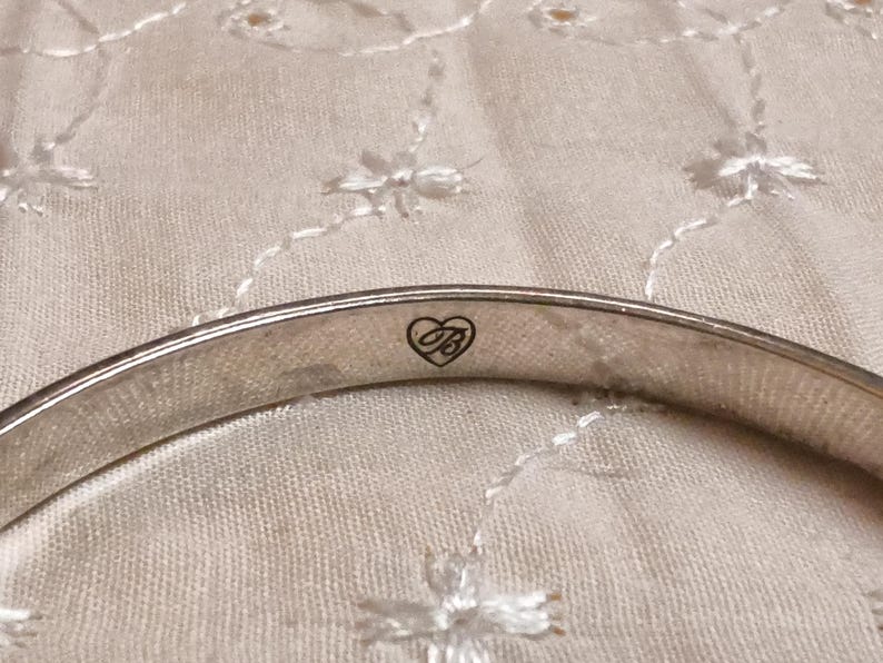 May include: Close-up of a silver-toned bangle bracelet. The bracelet has a smooth, polished surface and a heart-shaped logo with the letters "B" and "B" inside. The background is a cream-colored fabric with a floral pattern.