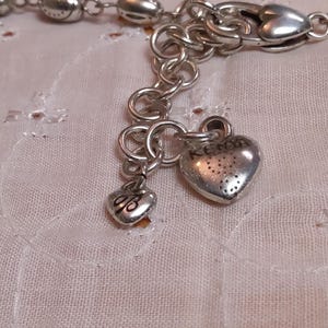 Op de afbeelding: Zilverkleurige armband met hartvormige bedels. De armband heeft een reeks schakels en decoratieve kralen. Twee hartvormige bedels hangen aan de armband, een met het woord "KENS" en de andere met de initialen "dB".