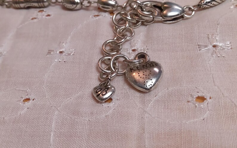 Op de afbeelding: Zilverkleurige armband met hartvormige bedels. De armband heeft een reeks schakels en decoratieve kralen. Twee hartvormige bedels hangen aan de armband, een met het woord "KENS" en de andere met de initialen "dB".