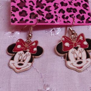 Pu&ograve; includere: Un paio di orecchini di Minnie Mouse. Ogni orecchino presenta un contorno dorato, testa e orecchie nere, viso bianco e fiocco rosso a pois bianchi. Gli orecchini pendono da ganci dorati.