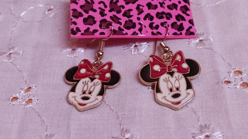 Pu&ograve; includere: Un paio di orecchini di Minnie Mouse. Ogni orecchino presenta un contorno dorato, testa e orecchie nere, viso bianco e fiocco rosso a pois bianchi. Gli orecchini pendono da ganci dorati.