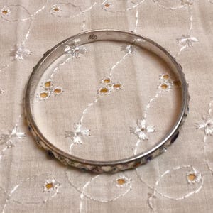 May include: A silver-toned bangle bracelet with colorful enamel detailing. The bracelet is circular and rests on a cream-colored fabric with embroidered floral patterns. The enamel features a variety of colors, adding a decorative touch to the jewelry.