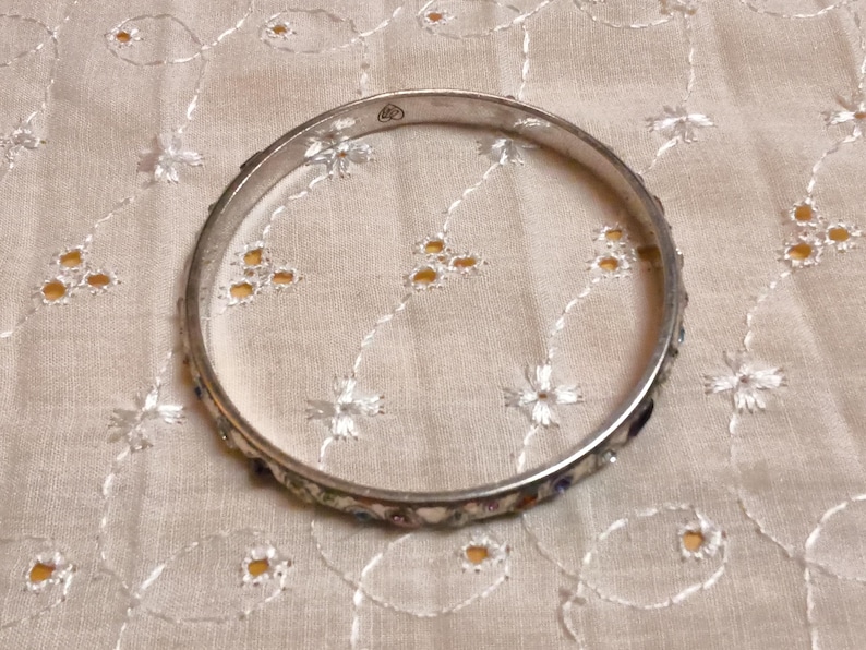 May include: A silver-toned bangle bracelet with colorful enamel detailing. The bracelet is circular and rests on a cream-colored fabric with embroidered floral patterns. The enamel features a variety of colors, adding a decorative touch to the jewelry.