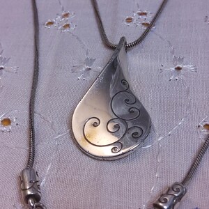 May include: A silver necklace with a teardrop-shaped pendant featuring a swirling design. The pendant is attached to a silver chain.