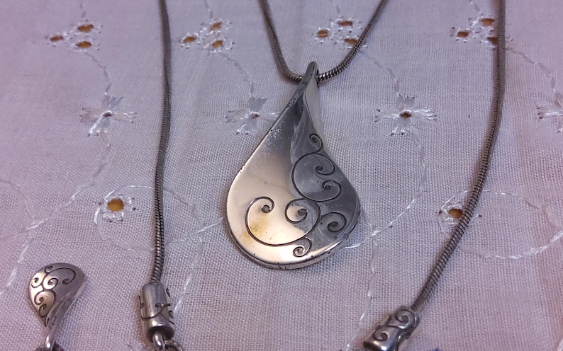 May include: A silver necklace with a teardrop-shaped pendant featuring a swirling design. The pendant is attached to a silver chain.