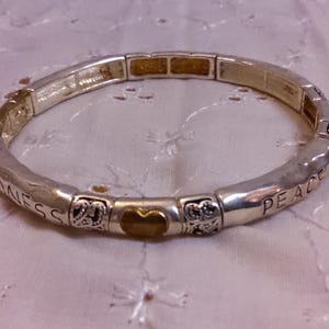 May include: A silver bracelet with a gold heart charm and the words "Peace" and "Goodness" engraved on it.