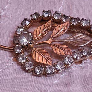 May include: Vintage leaf-shaped brooch with clear rhinestones and gold-toned metal. The brooch features a central cluster of leaves in gold and silver tones, surrounded by a ring of sparkling stones. A classic piece of jewelry.
