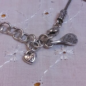 May include: Silver charm bracelet with a heart charm engraved with the letter "B" and a decorative charm with a swirl design.