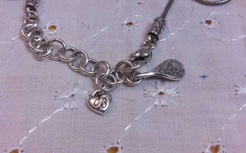 May include: Silver charm bracelet with a heart charm engraved with the letter "B" and a decorative charm with a swirl design.