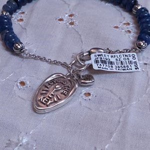Puede incluir: Una pulsera con cuentas azul oscuro y un dije plateado. El dije es de forma ovalada con las palabras "LOVE THE EARTH" grabadas. Tambi&eacute;n hay un peque&ntilde;o dije de coraz&oacute;n adjunto. La pulsera tiene una etiqueta adjunta con texto.