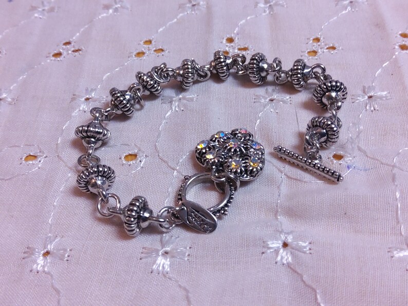 May include: A silver bracelet with a silver clasp and a silver charm with iridescent rhinestones. The bracelet is made of small, round beads with a textured surface.