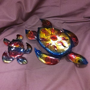 May include: Two metal sea turtles with a sun design on the shell. The larger turtle is blue, red, and yellow. The smaller turtle is red, blue, and yellow.