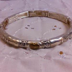 May include: A silver expandable bracelet with a gold heart charm and the words "Peace" and "I Love" engraved on the band.