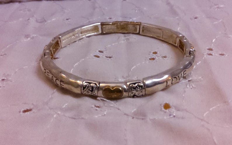May include: A silver expandable bracelet with a gold heart charm and the words "Peace" and "I Love" engraved on the band.