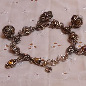 May include: Silver-toned charm bracelet with ornate, detailed links and charms. The bracelet features gold-toned accents and small, round, amber-colored stones. The clasp is gold-toned with a small heart charm. The bracelet is displayed on a white, patterned surface.