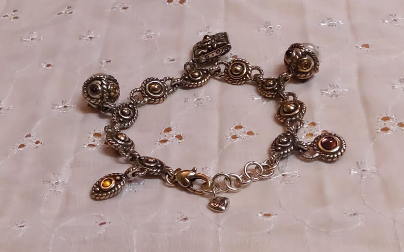 May include: Silver-toned charm bracelet with ornate, detailed links and charms. The bracelet features gold-toned accents and small, round, amber-colored stones. The clasp is gold-toned with a small heart charm. The bracelet is displayed on a white, patterned surface.