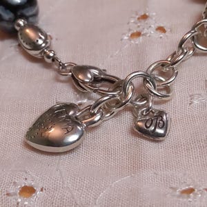 May include: Close-up of a silver bracelet with a heart-shaped charm engraved with the word "EMA". The bracelet features a small heart charm with the initials "OB". The bracelet is on a white, textured surface.