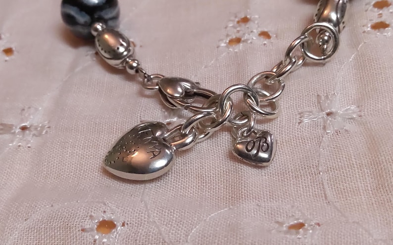 May include: Close-up of a silver bracelet with a heart-shaped charm engraved with the word "EMA". The bracelet features a small heart charm with the initials "OB". The bracelet is on a white, textured surface.