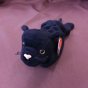 May include: Black plush toy cat with a red and white tag. The cat has a black nose, white whiskers, and brown eyes.