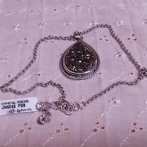Może przedstawiać: Srebrny naszyjnik z wisiorkiem w kształcie łzy wypełnionym ciemnymi, błyszczącymi kryształami. Wisiorek ma ozdobną obw&oacute;dkę. Naszyjnik jest prezentowany na jasnor&oacute;żowej, wzorzystej tkaninie. Dołączona jest etykieta z napisem "Crystal Rocks".