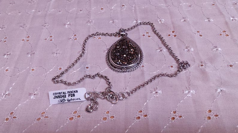 Może przedstawiać: Srebrny naszyjnik z wisiorkiem w kształcie łzy wypełnionym ciemnymi, błyszczącymi kryształami. Wisiorek ma ozdobną obw&oacute;dkę. Naszyjnik jest prezentowany na jasnor&oacute;żowej, wzorzystej tkaninie. Dołączona jest etykieta z napisem "Crystal Rocks".