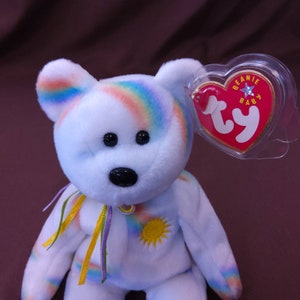 May include: White plush rainbow bear with a yellow sun on its chest and a Ty Beanie Baby tag attached to its ear. The tag is red with white lettering and a gold star.