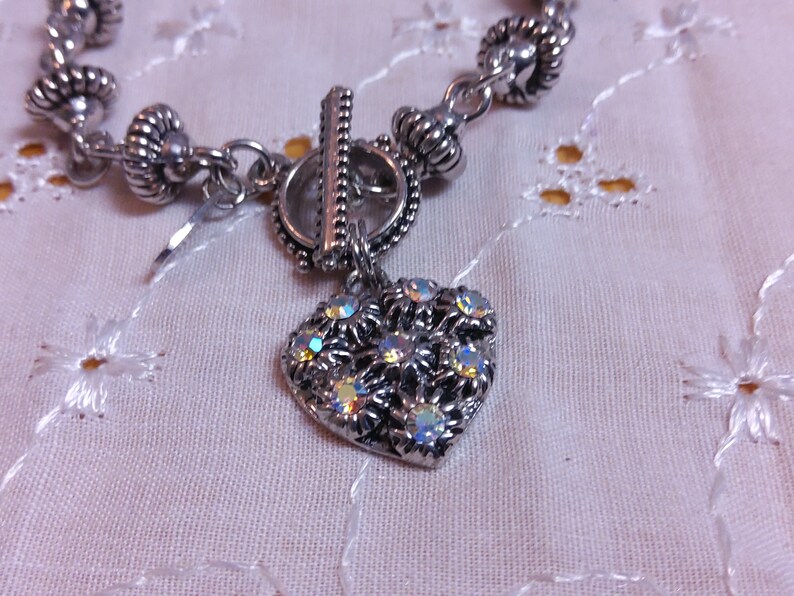 May include: A silver bracelet with a heart-shaped charm. The charm is decorated with iridescent rhinestones in a floral pattern.
