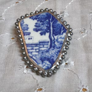May include: A brooch featuring a shard of blue and white ceramic pottery with a landscape scene of trees and a stone wall. The ceramic piece is encased in a silver-toned metal frame with a beaded edge, creating a shield-like shape.