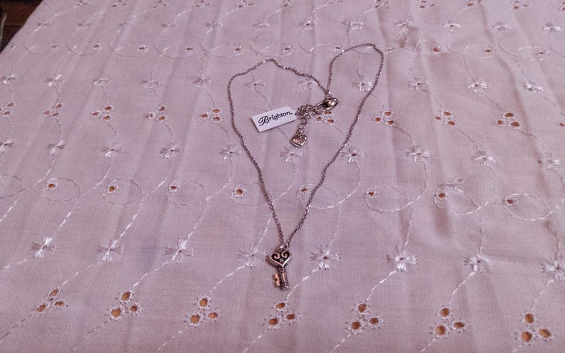 Op de afbeelding: Zilverkleurige ketting met een sleutelhanger. De sleutelhanger heeft een hartvormige bovenkant en een gedetailleerd sleutelontwerp. De ketting heeft een "Brighton"-label. Het ligt op een lichtroze, geborduurde stof. De ketting is ongeveer 45 cm lang.