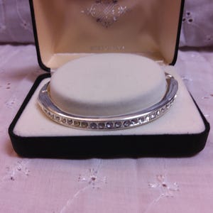 May include: A silver bangle bracelet with a row of clear rhinestones set in a silver band. The bracelet is in a black velvet box with a white satin lining.