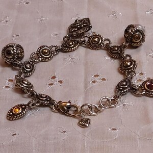 May include: Silver-toned charm bracelet with various decorative charms. The bracelet features a lobster clasp and a heart-shaped charm. Charms include round and oval shapes with gold and red accents. A decorative jewelry piece.