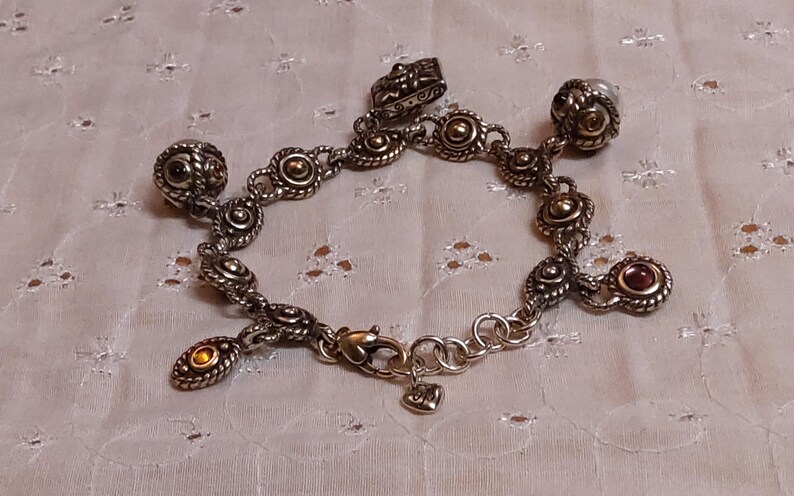 May include: Silver-toned charm bracelet with various decorative charms. The bracelet features a lobster clasp and a heart-shaped charm. Charms include round and oval shapes with gold and red accents. A decorative jewelry piece.