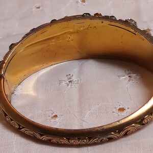 May include: A vintage gold-toned hinged bangle bracelet. The bracelet features an ornate, engraved design along the edges and is adorned with small, colorful gemstones. The interior has a warm, aged appearance, suggesting its antique nature.